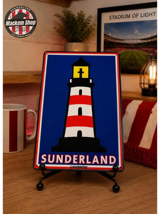 A rectangular metal-style sign on a small black stand, featuring a red-and-white striped lighthouse on a blue background with the word "SUNDERLAND" at the bottom. The sign is photographed on a wooden table in a cozy indoor setting with a mug, plant, framed stadium poster, and lantern visible in the background.