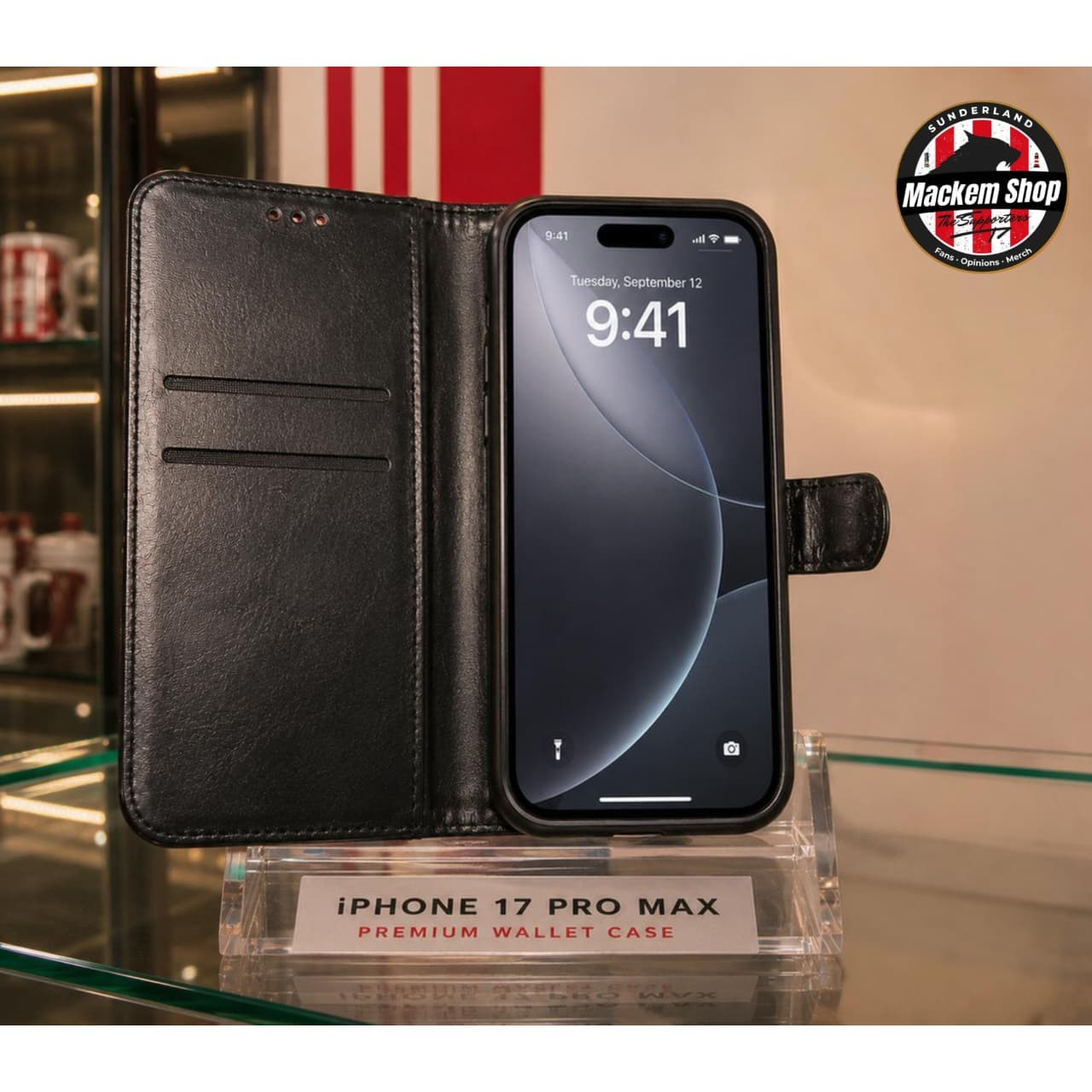 Black faux leather wallet case for iPhone 17 Pro Max, shown open with a phone inside on a glass display shelf. A circular 'Mackem Shop' logo is visible in the top right corner.