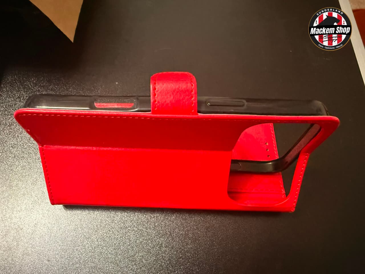 A bright red folio-style phone case with a kickstand, shown standing open on a dark surface. A circular 'Mackem Shop' logo watermark is visible in the top right corner.