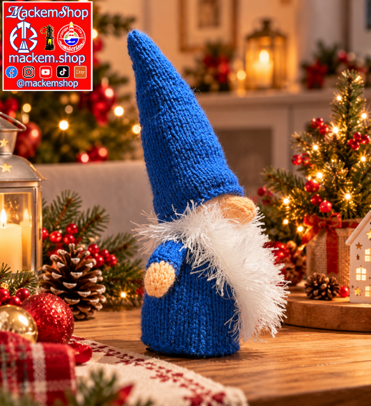 A blue knitted Gonk with a white fluffy beard and small beige nose, standing on a wooden table in a decorated holiday setting with candles, pinecones, and a lit Christmas tree. A red and white MackemShop logo overlay is visible in the top left corner.