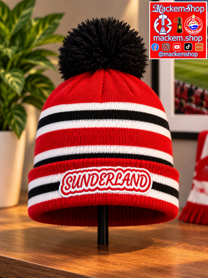 A red knit pom-pom beanie with black and white horizontal stripes and a white 'SUNDERLAND' patch on the front, displayed on a stand on a wooden surface with a plant in the background.