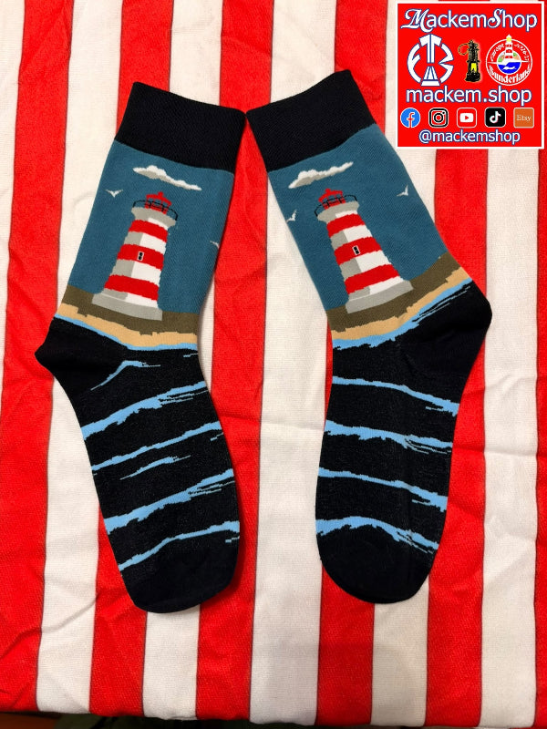 A pair of crew socks with a lighthouse and ocean design, laid flat on a red and white striped fabric background. The socks feature a red and white striped lighthouse on a blue-sky background with beige sand, and black and blue striped ocean waves below, and a MackemShop logo on top right corner.