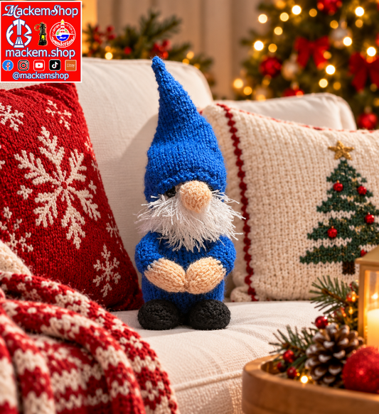 A small hand-knit gonk sits on a white sofa surrounded by holiday decor, including a red knit pillow with white snowflakes, a cream knit pillow with a Christmas tree design, a red knit throw blanket, and a blurred Christmas tree in the background.