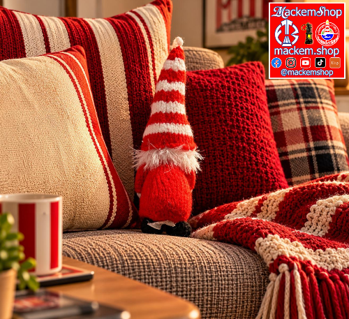A cozy living room scene with a small red-and-white striped gonk placed on a textured sofa among red and cream throw pillows and a red-and-cream knit blanket. A mug, phone, and small plant are visible on a table in the foreground.
