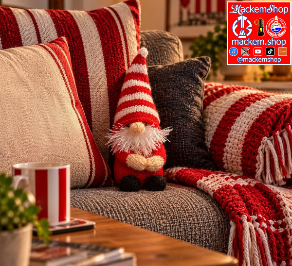 A festive red and white striped knit Gonk sits on a gray textured sofa surrounded by matching red and cream striped pillows and a red-and-white striped knit throw blanket. A coffee table and mug are visible in the foreground, and a MackemShop Logo appears in the top-right corner.