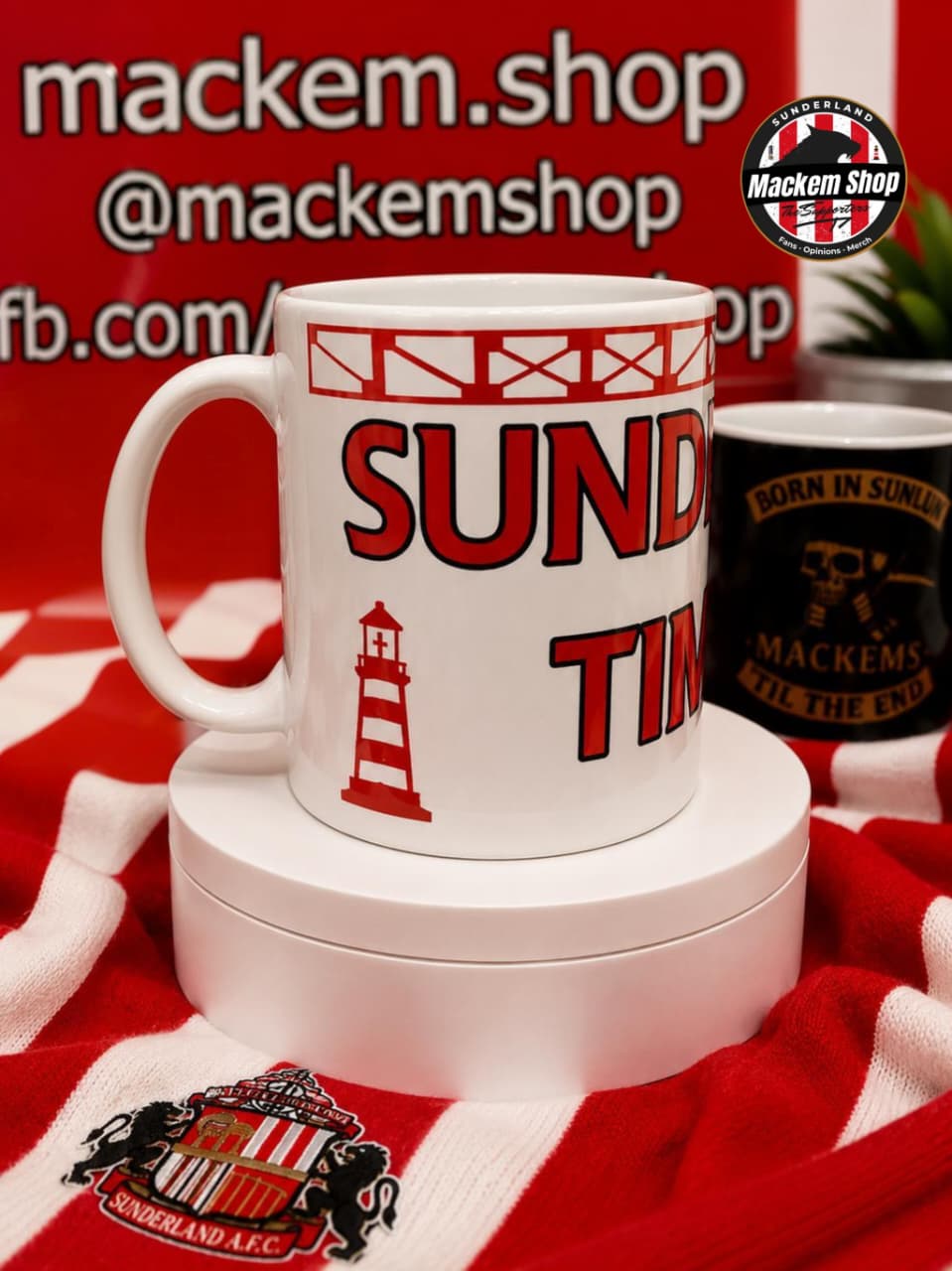 A white ceramic mug with red and black Sunderland AFC text and graphics, displayed on a white pedestal with a red and white striped scarf and a second black mug in the background. Prominent 'mackem.