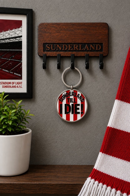 A Sunderland AFC themed keychain holder on a gray wall, featuring a wooden plaque with four black hooks and a round metal keychain charm that reads 'SUNDERLAND TILL I DIE!'. A framed stadium print, a small potted plant, and a red-and-white striped scarf are arranged around it.