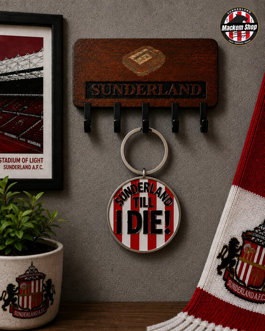 A Sunderland AFC themed keychain holder on a gray wall, featuring a wooden plaque with four black hooks and a round metal keychain charm that reads 'SUNDERLAND TILL I DIE!'. A framed stadium print, a small potted plant, and a red-and-white striped scarf are arranged around it.