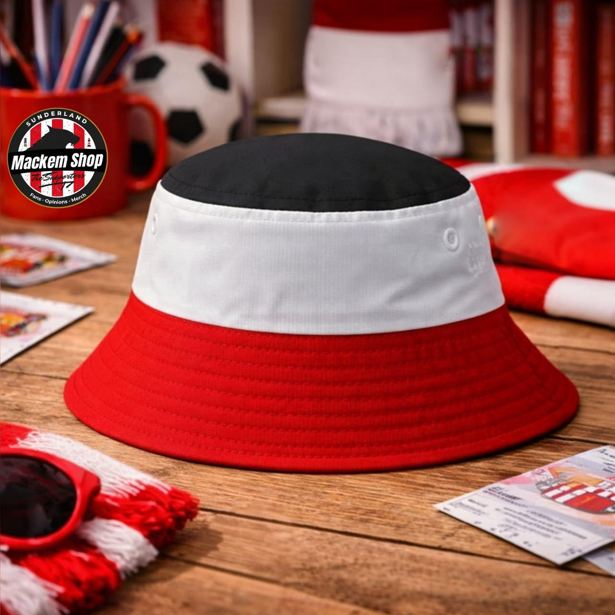 A bucket hat with a black top, white middle, and red brim, styled on a wooden table with a red and white scarf, sunglasses, and soccer-themed background props. A circular 'Mackem Shop' logo is visible in the top left corner.