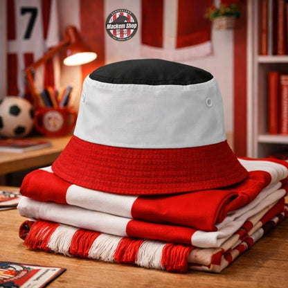 A red, white, and black bucket hat sits on top of folded red and white striped scarves on a wooden table. The background shows a cozy, blurred sports-themed room with a soccer ball, desk lamp, and bookshelves.