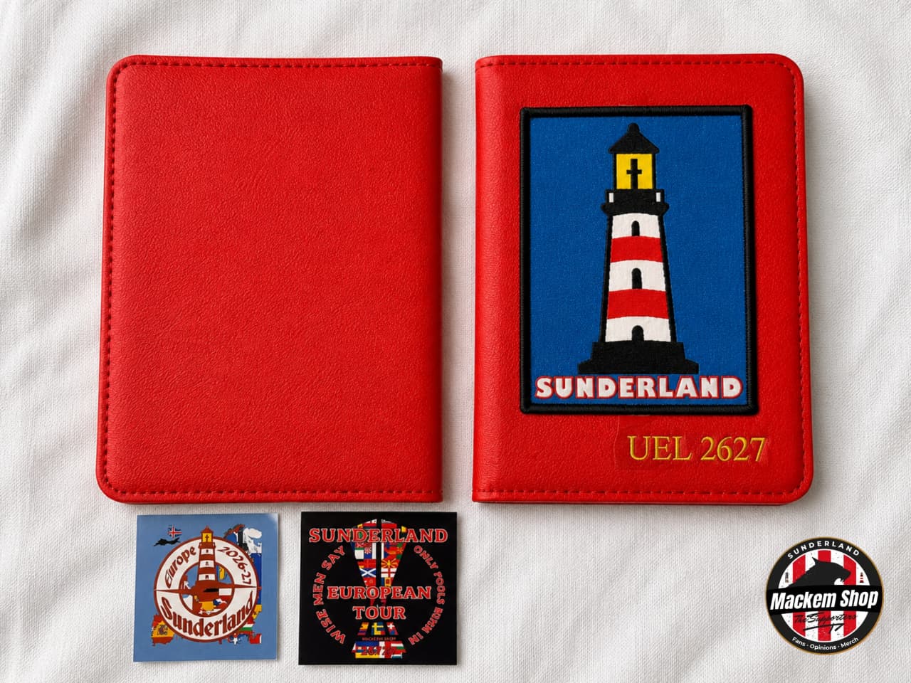 A red faux-leather passport cover shown closed and open, with a printed Sunderland lighthouse patch on the inside cover. Two small printed inserts are placed below, and a circular 'Macken Shop' logo appears in the lower right corner, all laid on a white fabric background.