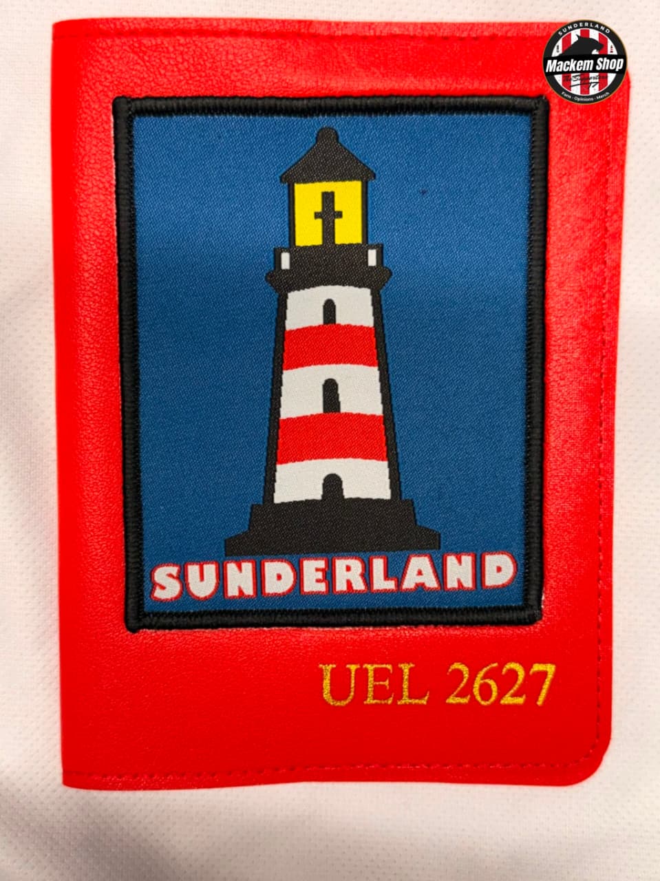 A red leather Passport cover featuring an embroidered patch of a red and white striped lighthouse on a blue background with the word 'SUNDERLAND' and the text 'UEL 2627' below. A circular 'Mackem Shop' logo is visible in the top right corner.