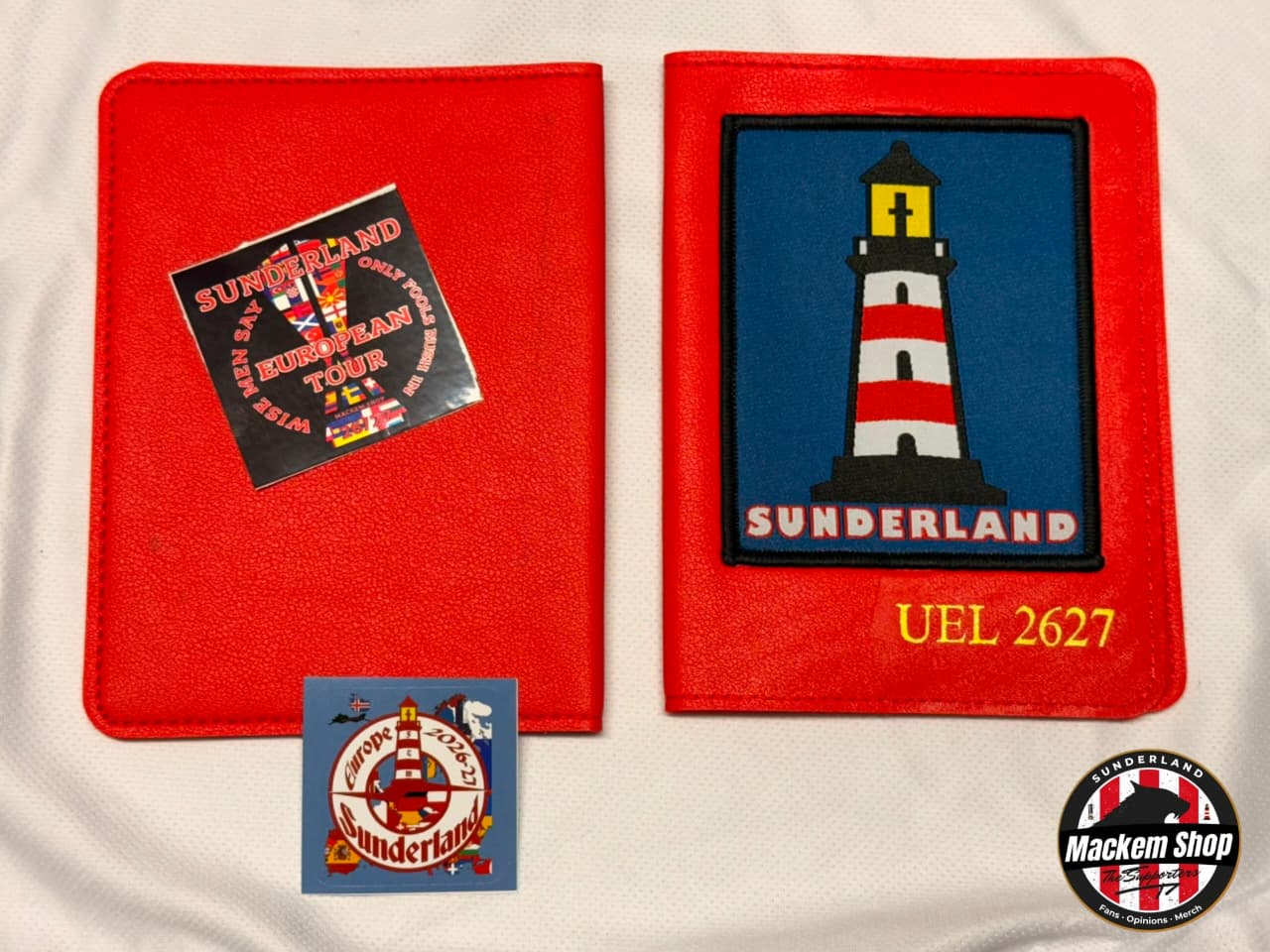 A red embroidered Passport cover set displayed on a white fabric background. The set includes a red rectangular patch with a black Sunderland tour sticker, a larger red patch featuring a red-and-white striped lighthouse on a blue background with 'SUNDERLAND' and 'UEL 2627' in yellow, and a small blue circular patch.