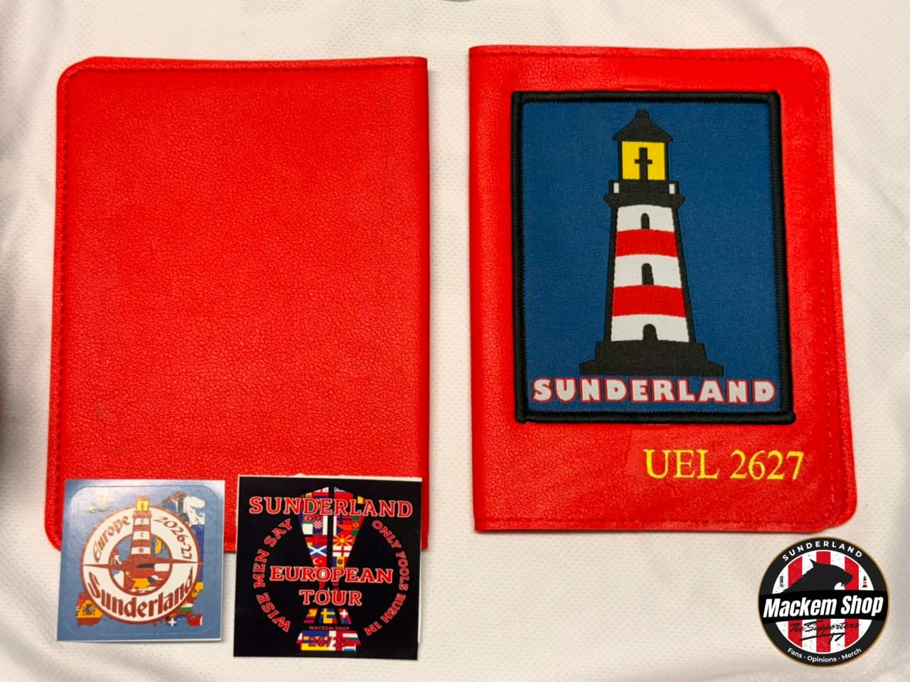A red fabric Souvenir Passport cover shown from both front and back, featuring a lighthouse patch on the front with 'SUNDERLAND' and 'UEL 2627' text, plus two small, attached stickers, photographed on a white fabric background.