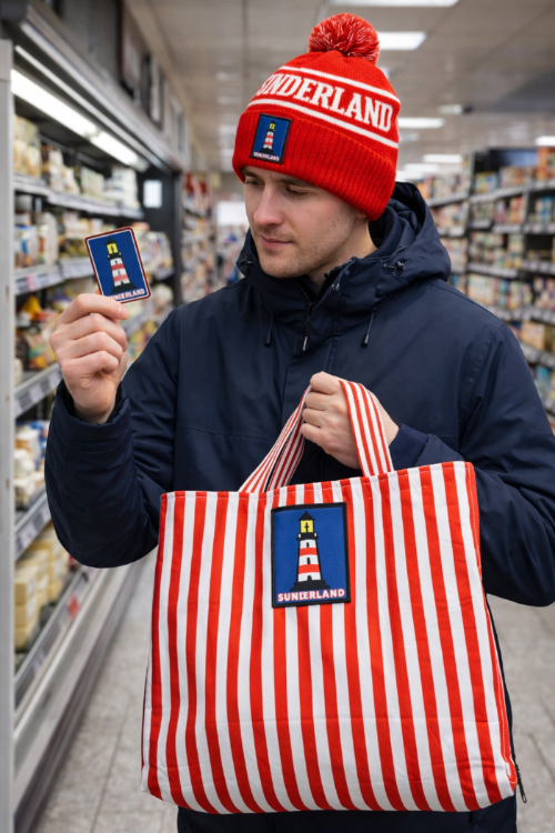 Person wearing Sunderland Bobble hat while holding Sunderland Lighthouse fridge magnet in one hand and Handmade Sunderland Tote bag in other hand.