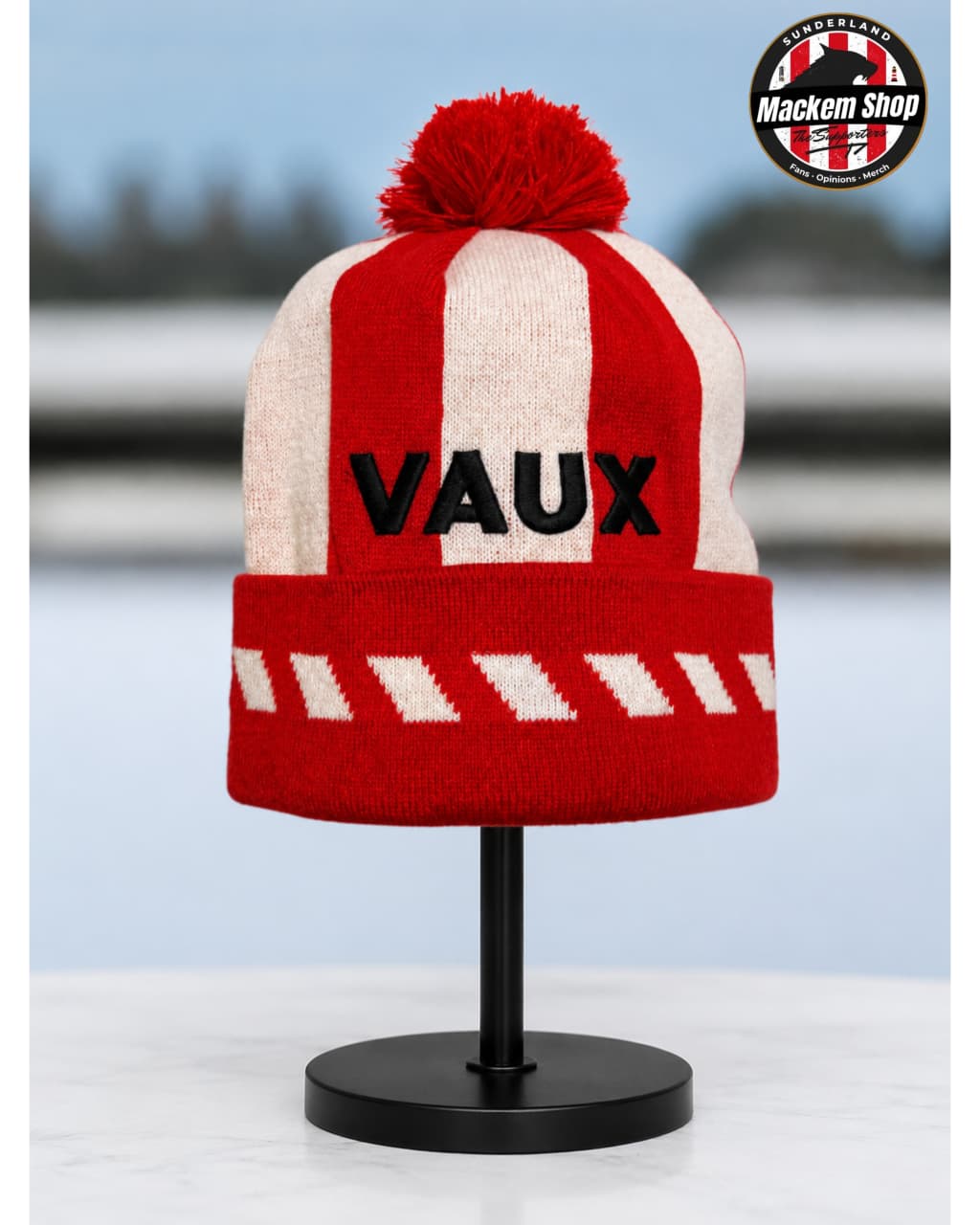 A red and off-white striped knit beanie with a red pom-pom on top and the word 'VAUX' in black across the front, displayed on a black stand against a blurred outdoor background. A circular 'Mackem Shop' logo appears in the top right corner.