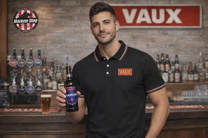 Man holding a Vaux beer bottle in a bar while wearing VAUX polo shirt in a setting with Vaux branding visible.