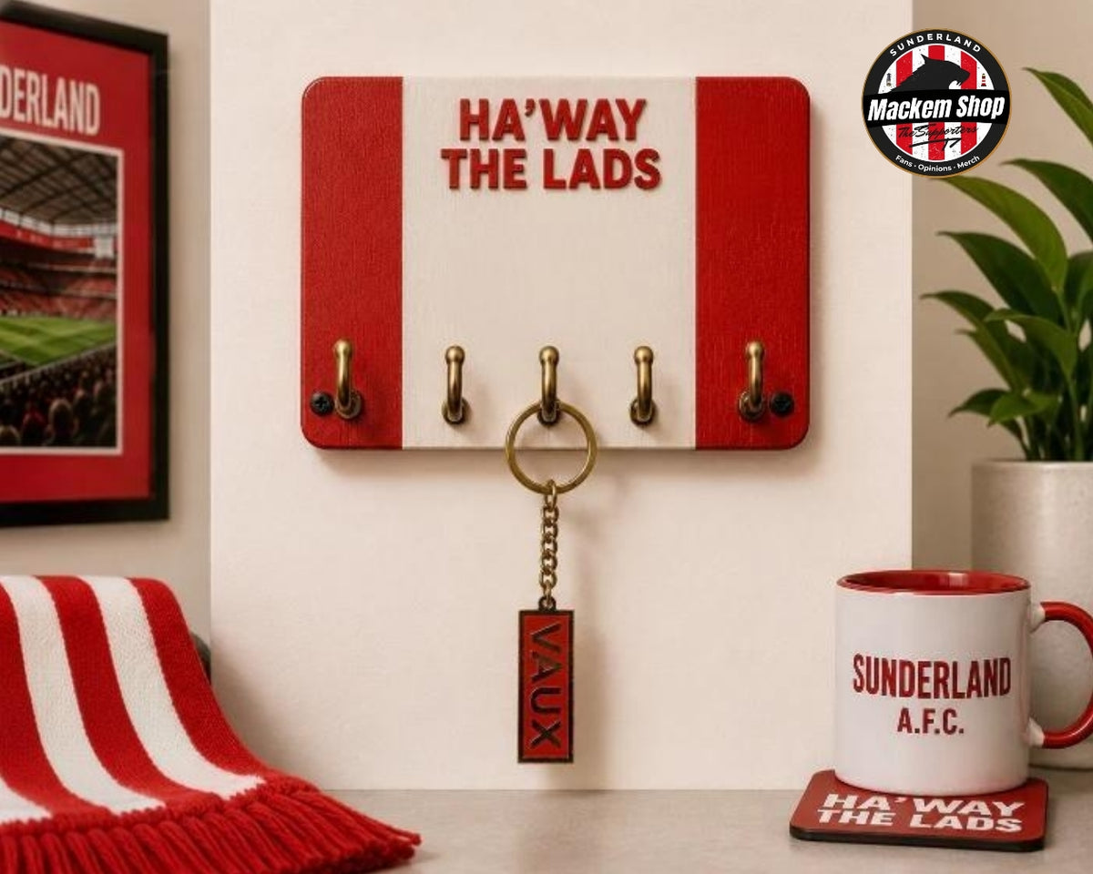 VAUX name Keyring on a red and white wooden key holder with four brass hooks and the words 'HA'WAY THE LADS' in red, mounted on a wall. A red and white striped scarf, a framed Sunderland picture, a potted plant, and a white mug with red trim are arranged around it.