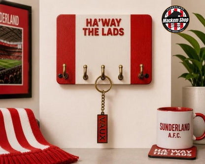 VAUX name Keyring on a red and white wooden key holder with four brass hooks and the words 'HA'WAY THE LADS' in red, mounted on a wall. A red and white striped scarf, a framed Sunderland picture, a potted plant, and a white mug with red trim are arranged around it.