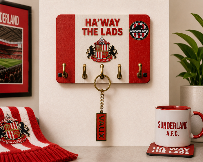 Wall hook with 'Ha' Way the Lads' text, mug, "VAUX" Keychain and scarf on a table with a plant in the background.
