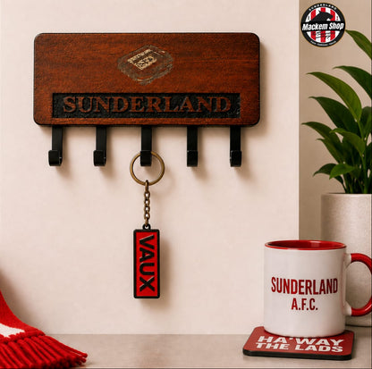A wooden Sunderland AFC wall key holder with four black hooks, shown mounted on a beige wall in a styled room scene. A red and white Sunderland scarf is draped on the left, a framed Sunderland picture is partially visible, and a white Sunderland A.