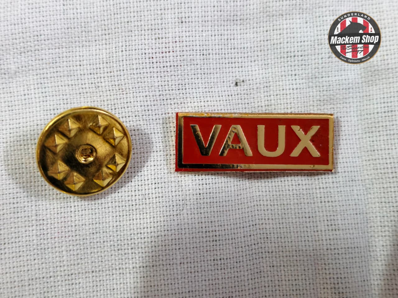 Gold metal button and red 'VAUX' badge on a textured white background with 'Mackem Shop' logo.
