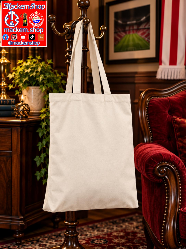 A plain off-white canvas tote bag hanging from a dark wooden coat rack in a furnished room with a red velvet armchair, wood furniture, and framed wall art. A red and white MackemShop logo overlay appears in the top-left corner.