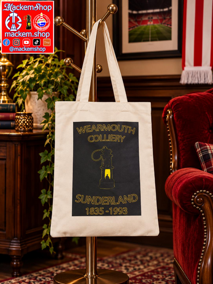 A tote bag hanging on a brass coat rack in a vintage-style room. The tote bag is off-white with a large black graphic panel featuring gold text that reads 'WEARMOUTH COLLIERY' and 'SUNDERLAND 1835-1993' with a lantern illustration. A MackemShop Logo is visible on top left corner.