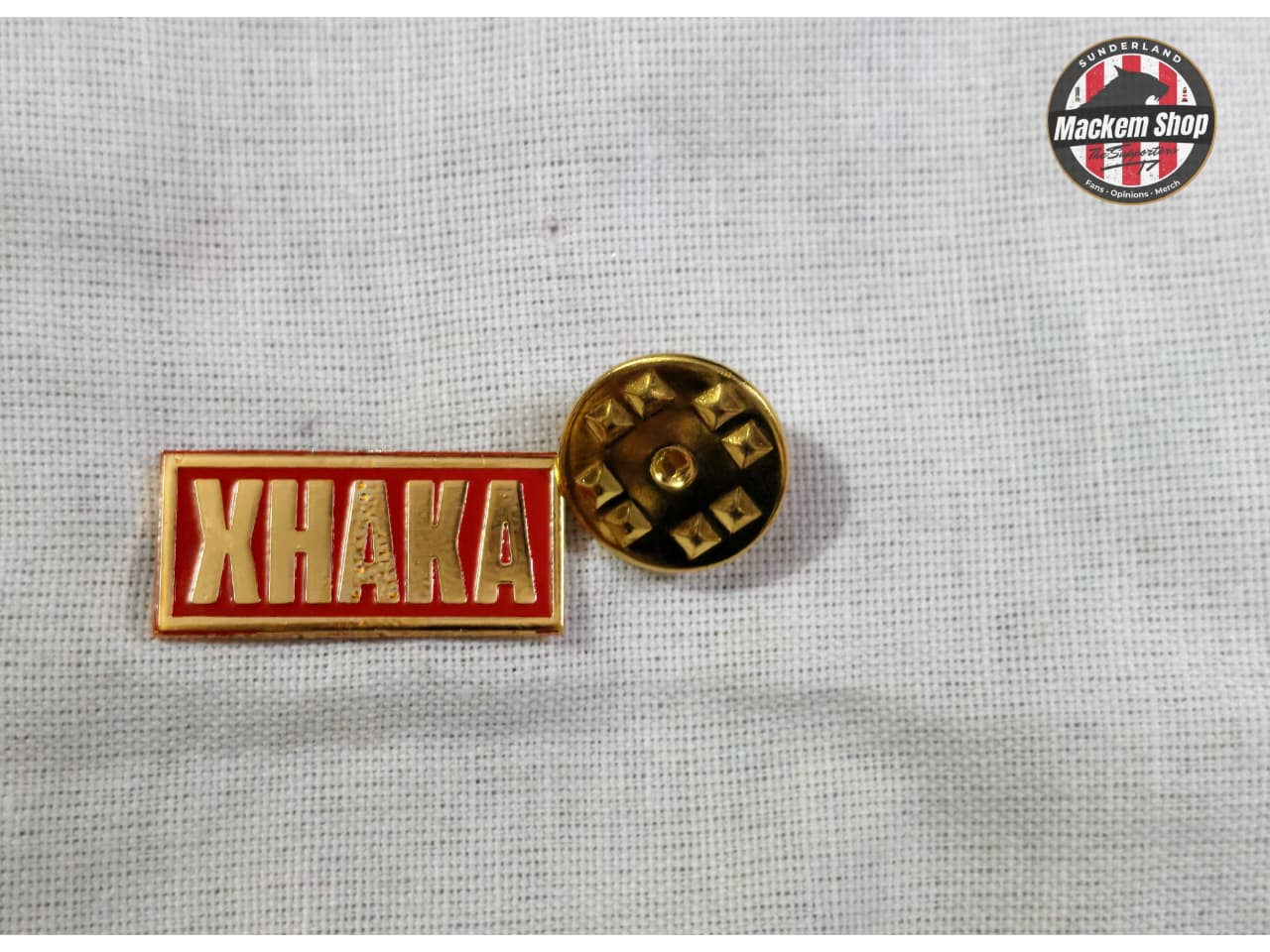 Two pins, one red with 'XHAKA' text and one gold, on a gray background.