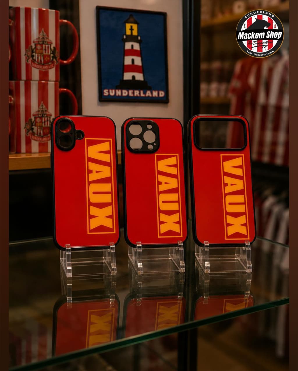 Three red phone cases with 'VAUX' branding on clear stands, with a blurred background featuring a lighthouse painting and a red and white striped object.